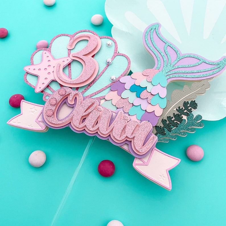 May include: A layered mermaid-themed cake topper with the name "Olivia" and the number "3". The topper features a pink glitter finish, a starfish, and a mermaid tail in shades of purple and blue. Decorative elements include small pearls and a banner.