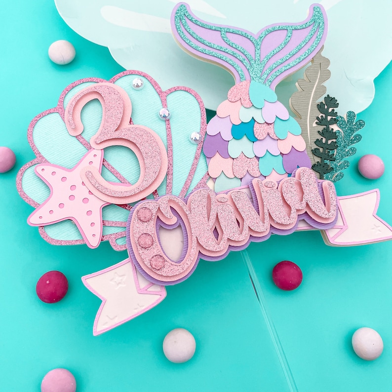 May include: A mermaid-themed cake topper with the name "Olivia" and the number "3". The topper features a mermaid tail, a seashell, and a starfish, all in pastel colors with glitter accents. The background is a light blue.