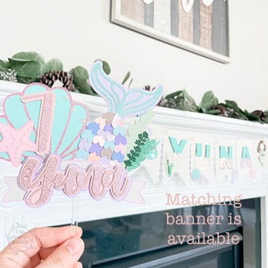 May include: A mermaid-themed cake topper with the name "Yuna" and the number "7" in pastel colors. The topper features a mermaid tail, seashells, and a banner. A matching banner is available.