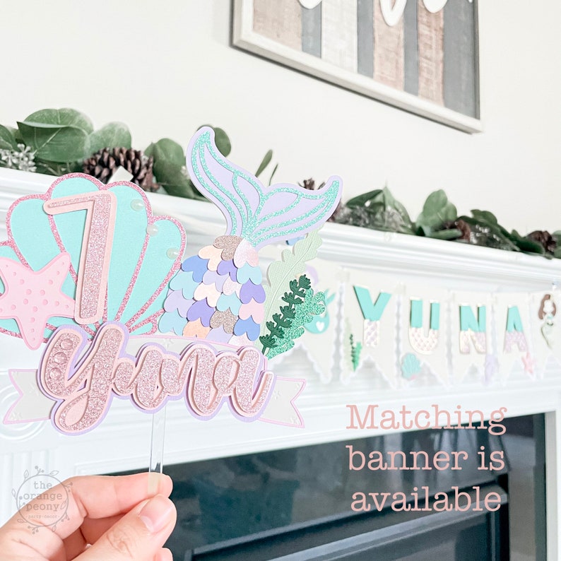 May include: A mermaid-themed cake topper with the name "Yuna" and the number "7" in pastel colors. The topper features a mermaid tail, seashells, and a banner. A matching banner is available.