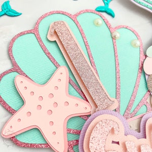 May include: A layered paper craft with a mermaid theme. Features a light blue seashell shape with a pink glitter outline, a pink starfish, and a glittery number one. Decorative pearls and mermaid tail accents complete the design.