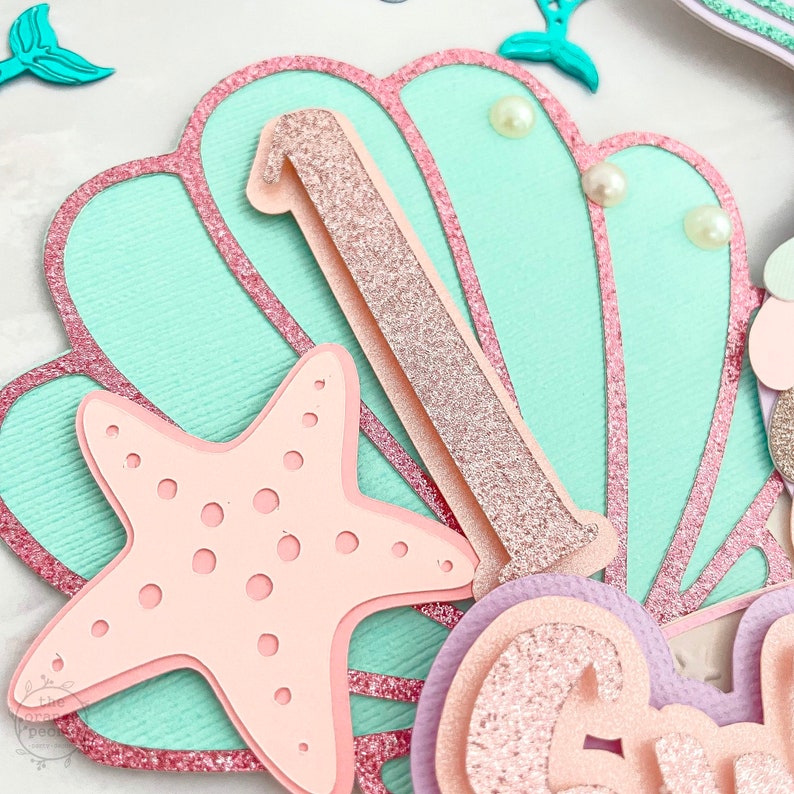 May include: A layered paper craft with a mermaid theme. Features a light blue seashell shape with a pink glitter outline, a pink starfish, and a glittery number one. Decorative pearls and mermaid tail accents complete the design.