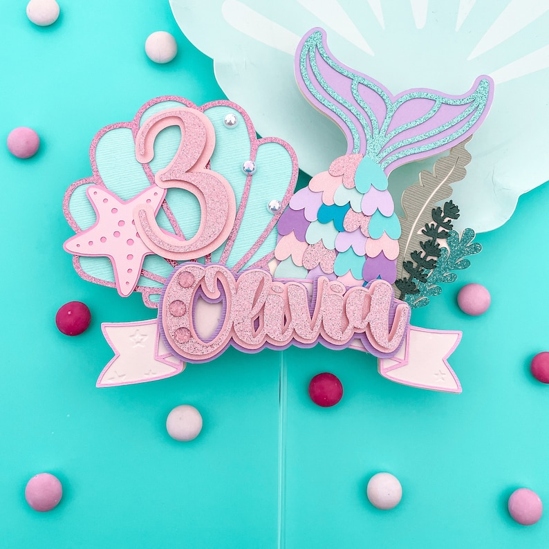 May include: A mermaid-themed cake topper with the name "Olivia" and the number "3". The topper features a mermaid tail, a seashell, and a starfish, all in shades of pink, purple, and teal. Decorative elements include glitter and faux gems.