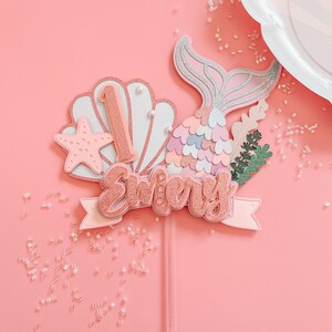 May include: A pink and glittery mermaid-themed cake topper with the name "Emery" and the number "1". The topper includes a mermaid tail, seashell, starfish, and decorative greenery. The background is a solid pink color.