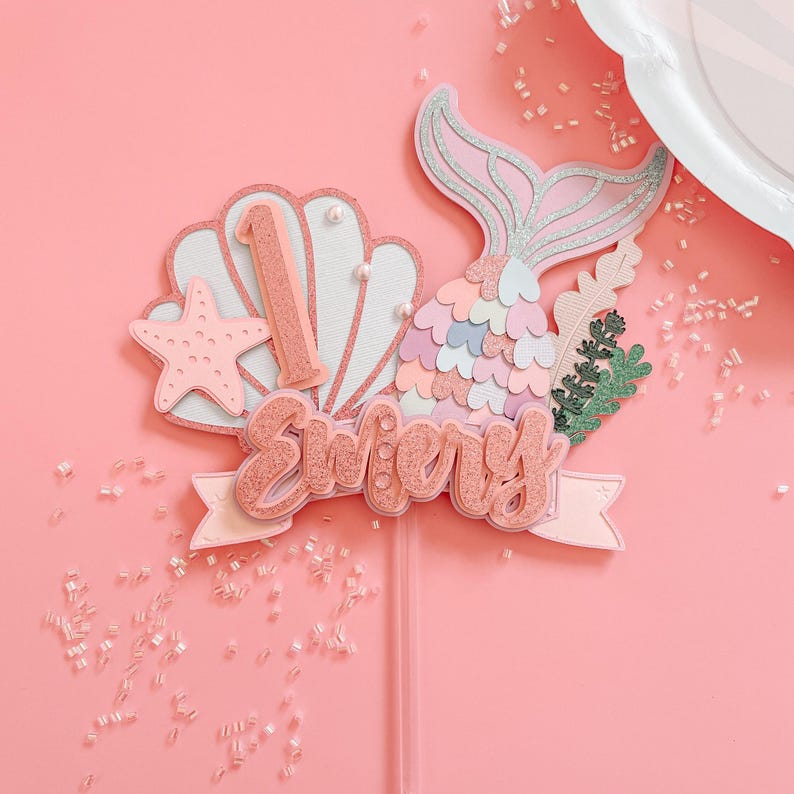 May include: A pink and glittery mermaid-themed cake topper with the name "Emery" and the number "1". The topper includes a mermaid tail, seashell, starfish, and decorative greenery. The background is a solid pink color.