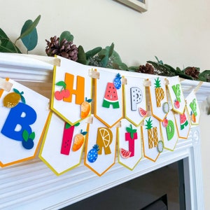 Può includere: Uno striscione di buon compleanno colorato con disegni di frutta e anguria. Ogni lettera è decorata con un frutto o una verdura diverso.