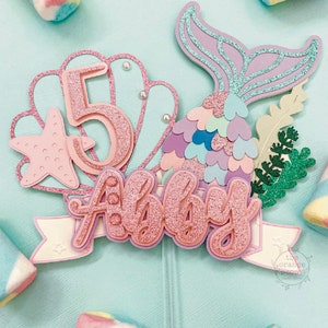 May include: A mermaid-themed cake topper with the name "Abby" and the number "5" in glittery pink. The design includes a mermaid tail, starfish, and seashell elements, all on a light blue background. Marshmallows surround the topper.