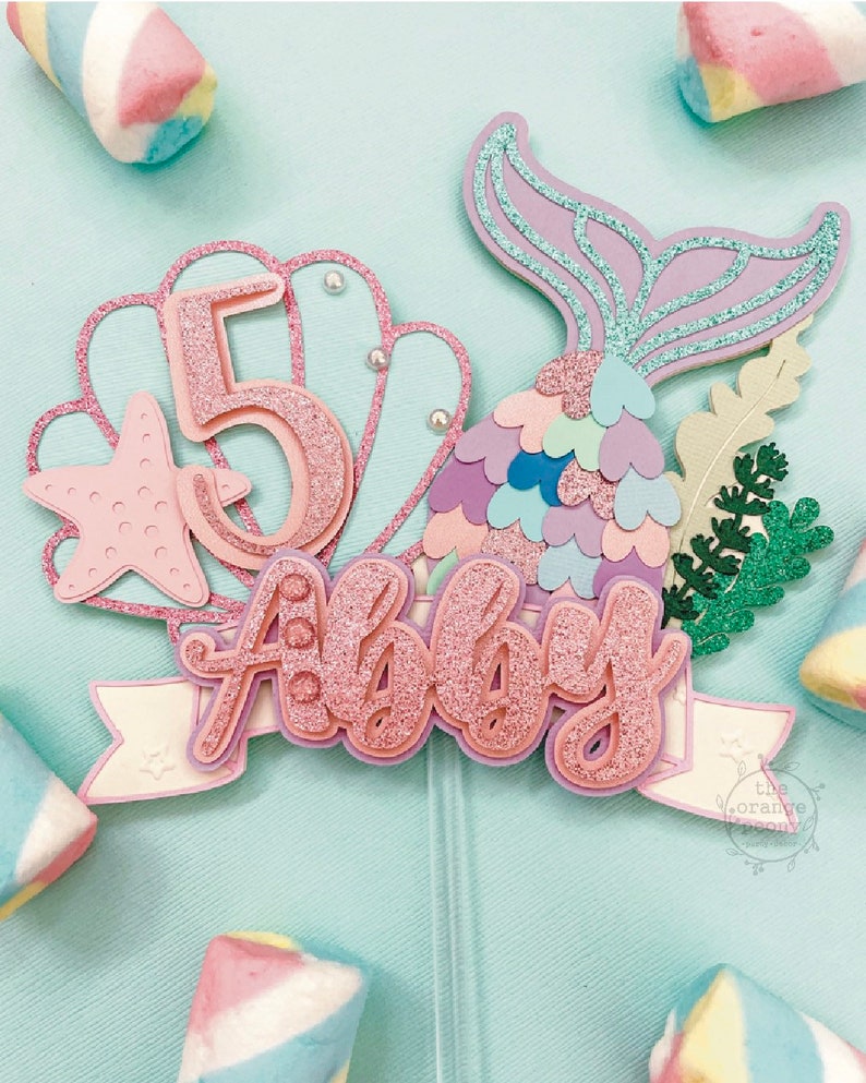 May include: A mermaid-themed cake topper with the name "Abby" and the number "5" in glittery pink. The design includes a mermaid tail, starfish, and seashell elements, all on a light blue background. Marshmallows surround the topper.