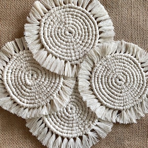 Puede incluir: Cuatro posavasos circulares de macramé color crema, con un diseño en espiral y bordes con flecos. Los posavasos están dispuestos sobre una superficie de arpillera marrón texturizada. Los posavasos son de color neutro y complementan cualquier decoración.