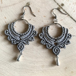 May include: A pair of gray macrame earrings with silver-colored metal accents. The earrings feature a half-circle design with intricate knotting and small silver beads. Each earring has a teardrop-shaped charm hanging from the bottom and a silver hook.