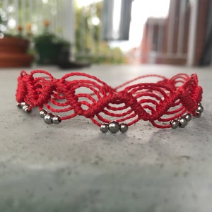 以下が含まれることがあります： 赤い紐のブレスレットに銀色のビーズが波模様に編み込まれている