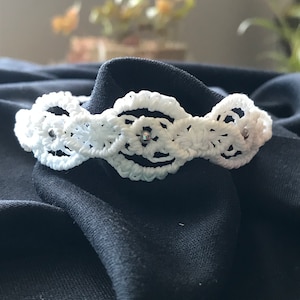 Peut inclure: Bracelet en crochet blanc avec des perles argentées. Le bracelet est sur un fond de tissu noir.