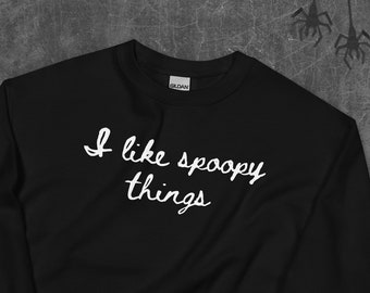May include: Black sweatshirt with the phrase "I like spoopy things" in white script. The sweatshirt is a crew neck style. The image has Halloween-themed decorations, including spider silhouettes and a sign that says "Happy Halloween".