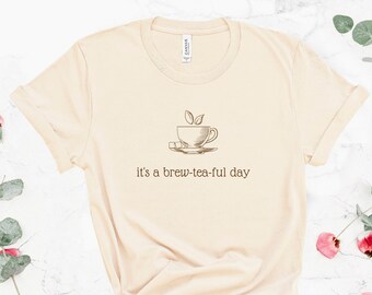 Puede incluir: Camiseta color crema con un gráfico de una taza de té y platillo, con vapor y dos hojas. El texto "it's a brew-tea-ful day" está impreso debajo. La camiseta está anudada en la parte inferior.