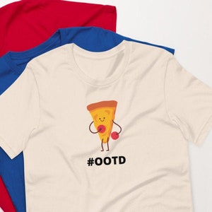 Puede incluir: Tres camisetas en colores rojo, azul y crema. La camiseta color crema presenta una porción de pizza de dibujos animados con guantes de boxeo y el texto "#OOTD". Las camisetas están hechas de un material suave.