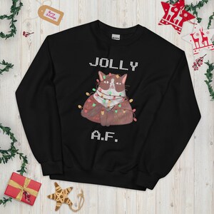 May include: Black sweatshirt with a graphic of a grumpy cat wrapped in Christmas lights. The text "JOLLY A.F." is displayed in white, pixelated font. Festive holiday apparel.