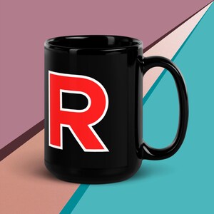 Peut inclure: Mug en céramique noire avec un grand "R" rouge et blanc. La lettre est en gras et sans empattement. La tasse a une anse incurvée et une finition brillante. Le fond est un mélange de formes géométriques roses, turquoise et blanches.