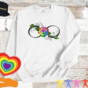 May include: White sweatshirt featuring a rainbow rose within a black infinity symbol. The rose has vibrant colors, and the design includes green leaves. A black bucket hat with a rainbow stripe is in the upper left corner.