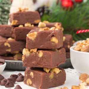 May include: A stack of chocolate fudge squares topped with walnuts. The fudge is a rich brown color, and the walnuts are a light tan. Chocolate chips and a bowl of walnuts are in the background. Festive holiday theme.