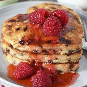 May include: A stack of heart-shaped pancakes with chocolate chips, topped with fresh raspberries and a vibrant red sauce. The pancakes are on a white plate, creating a visually appealing breakfast or dessert.