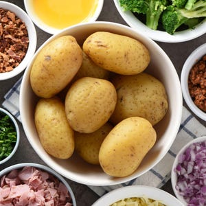May include: Overhead shot of a white bowl filled with golden-brown potatoes, surrounded by small bowls of ingredients. These include chopped broccoli, red onions, bacon bits, chives, and a bowl of what appears to be melted butter or oil. The ingredients are arranged on a gray surface.