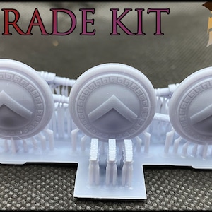 May include: A set of three 3D printed white plastic Spartan shields with a raised design. The shields are on a white plastic base with the text "UPGRADE KIT" and "DYNAMIC PRINTS STUDIO" above the shields.