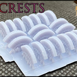 May include: A sheet of 20 white plastic crests, each with a detailed design, arranged in rows on a clear plastic sheet.  The sheet is labeled "20X CRESTS" in red and white text.