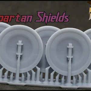 K&ouml;nnte beinhalten: F&uuml;nf graue, 3D-gedruckte Spartaner-Schilde mit einem erhabenen Design. Die Schilde sind an einer grauen Kunststoffbasis befestigt. Der Text "5 x Spartan Shields" ist oben im Bild sichtbar.