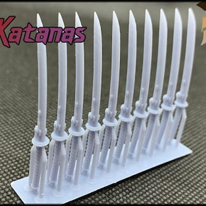 May include: A set of ten white plastic katana swords, arranged in a row on a white plastic base. The swords are all the same size and shape, and they have a slightly curved blade. The text "10x Katanas" is visible in the image.