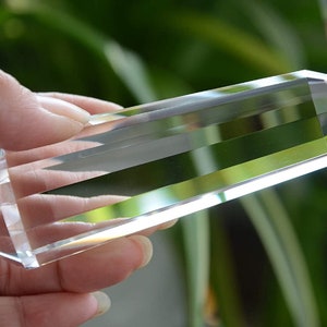 May include: A clear quartz crystal point, also known as a crystal wand, is held in a hand. The crystal is long and slender with a pointed tip. It is a popular tool for meditation and energy healing.