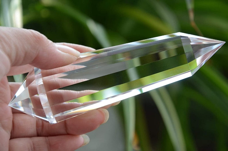 May include: A clear quartz crystal point, also known as a crystal wand, is held in a hand. The crystal is long and slender with a pointed tip. It is a popular tool for meditation and energy healing.