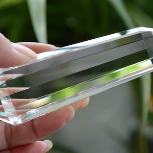 May include: A clear quartz crystal point, also known as a double terminated crystal, with a smooth, polished surface. The crystal is held in a hand, with a blurred green background.