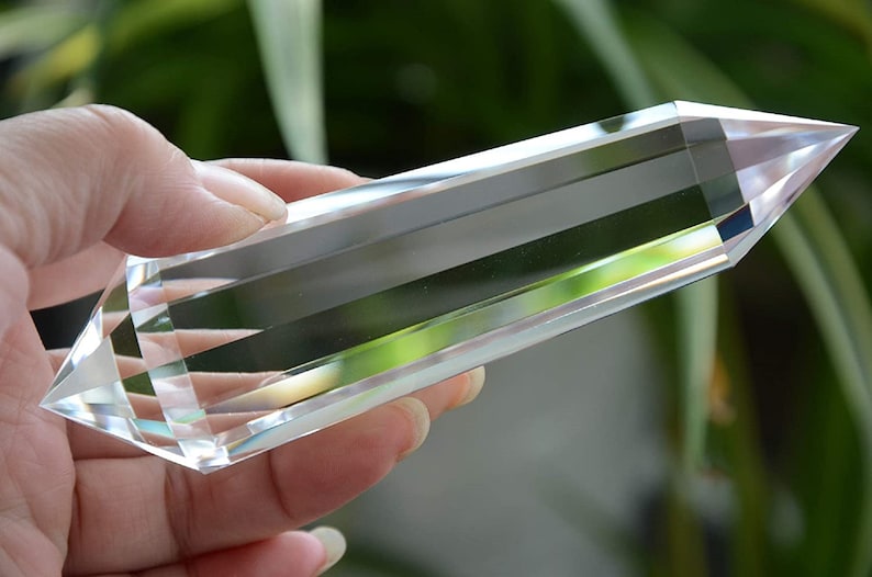 May include: A clear quartz crystal point, also known as a double terminated crystal, with a smooth, polished surface. The crystal is held in a hand, with a blurred green background.