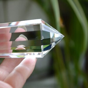 May include: A clear quartz crystal point with multiple facets, held in a hand against a blurred green background.