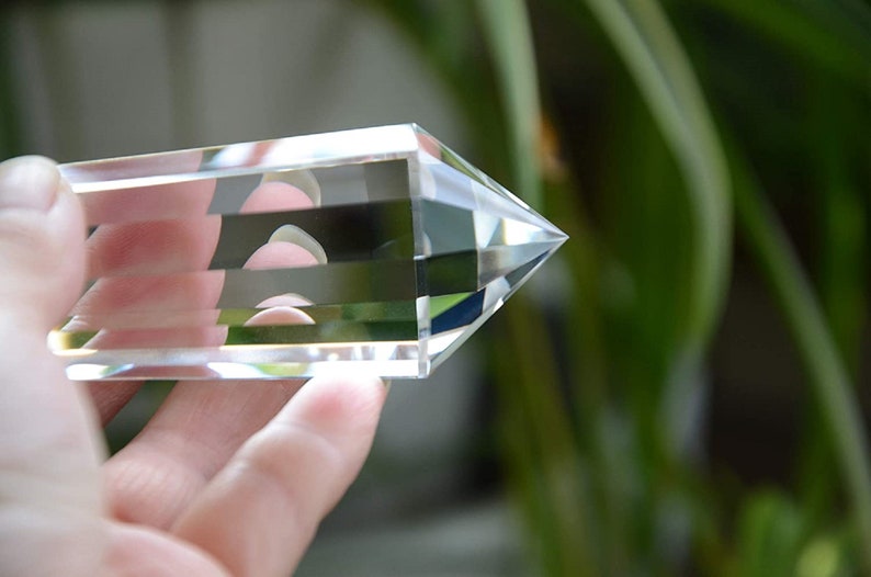 May include: A clear quartz crystal point with multiple facets, held in a hand against a blurred green background.