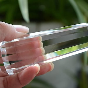 Puede incluir: Una varita de cristal clara y facetada, sostenida en una mano. El cristal es un punto de doble terminación, con múltiples facetas anguladas que crean un efecto brillante. El fondo es un follaje verde borroso.