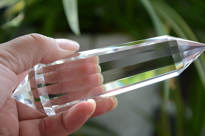 May include: A clear quartz crystal point, also known as a double terminated crystal, is held in a hand. The crystal is long and slender with a pointed tip at each end. The crystal is clear and transparent, and the light reflects off of its facets.
