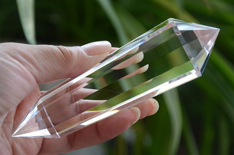 May include: A clear quartz crystal point, also known as a crystal wand, is held in a hand. The crystal is long and slender with a pointed tip. It is a popular tool for healing and meditation.