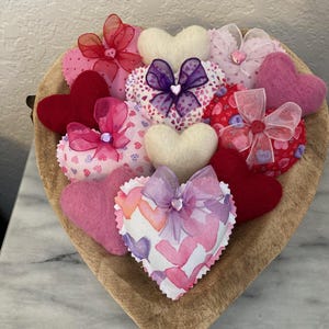 May include: A wooden heart-shaped bowl filled with various heart-shaped decorations. The hearts are made of felt and fabric in shades of red, pink, and white, some adorned with bows. The bowl sits on a marble surface.