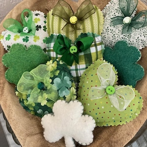 May include: A wooden bowl filled with St. Patrick's Day decorations. The decorations include heart-shaped and shamrock-shaped fabric ornaments in various shades of green, adorned with bows and buttons. A white felt shamrock is also present.