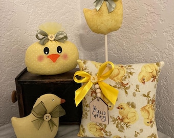 May include: Three yellow chick decorations with green bows. One chick is standing on a stick, another is sitting on a pillow, and the third is sitting on a wooden box. The pillow has a floral pattern with yellow chicks and the text "Hello Spring".