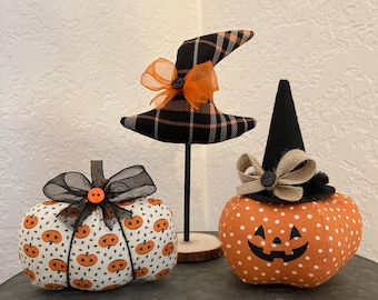 May include: Three Halloween-themed decorations: a witch hat on a stand, and two fabric pumpkins. The pumpkins are orange and white, with jack-o'-lantern faces and bows. The witch hat is black and orange plaid with an orange bow.