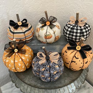 May include: Six fabric pumpkins with wooden stems and decorative bows. The pumpkins feature various Halloween-themed patterns, including stripes, ghosts, bats, and spiderwebs. Each pumpkin has a button accent, and the bows are in black, orange, and tan.