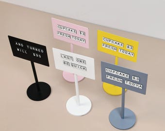 May include: Five standing metal signs in black, pink, white, yellow, and blue. Each sign features a rectangular board with white tiles displaying text: AND TURNED MILL $90, CUPCAKE $3 FRESH TODAY, LAST ONE BE QUICK, and CUPCAKE $3 FRESH TODYA.