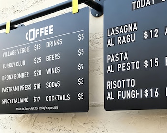 May include: Two black rectangular menu boards with white text and prices, suspended from a black metal bar with yellow clips. The left board lists coffee, sandwiches, and drinks with prices. The right board displays daily specials like lasagna, pasta, and risotto.