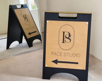 May include: A black A-frame sidewalk sign with a tan rectangular insert featuring the black text PACE STUDIO, a stylized PS logo inside an oval, and a left-pointing arrow. The sign has a gold-toned top handle and is reflected in a mirror.