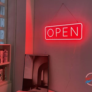 以下が含まれることがあります： 白い天井から吊り下げられた、白い文字で「OPEN」と書かれた赤いネオンサイン。サインは、本棚と額縁に入った絵画のある部屋にある。