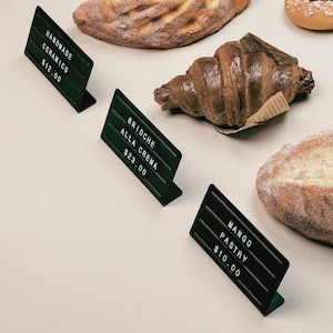 May include: Three dark green L-shaped metal display stands with white plastic letters and numbers. The signs read HANDMADE CERAMICS $12.00, BRIOCHE ALLA CREMA $23.00, and MANGO PASTRY $10.00, positioned next to various baked goods like bread and a croissant.