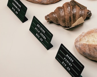 May include: Three dark green L-shaped metal display stands with white plastic letters and numbers. The signs read HANDMADE CERAMICS $12.00, BRIOCHE ALLA CREMA $23.00, and MANGO PASTRY $10.00, positioned next to various baked goods like bread and a croissant.
