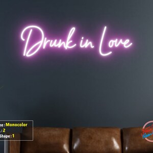 May include: Pink neon sign with the text "Drunk in Love" on a dark gray wall. The sign is mounted above a brown leather couch.
